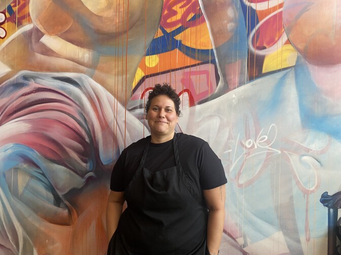 A woman with light, dark skin wearing a black apron and a black T-shirt underneath leans against a wall containing a large graffiti mural that covers the entire background. She has short, curly hair and is smiling with her hands inside her pockets. 
