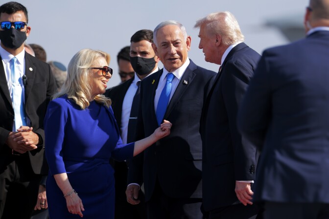 Two older white men in suits speak on a tarmac surrounded by security officials and a blonde woman in a bright blue dress.