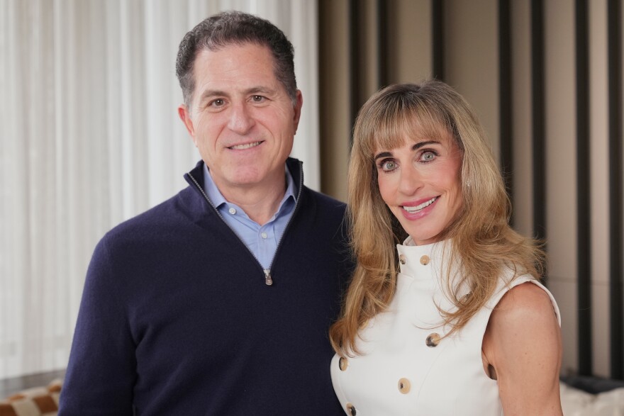Michael Dell, a man with light skin tone, wearing a navy blue quarter zipped sweater, poses for a photo with Susan Dell, a woman with light skin tone, wearing a cream white dress with gold buttons on it.