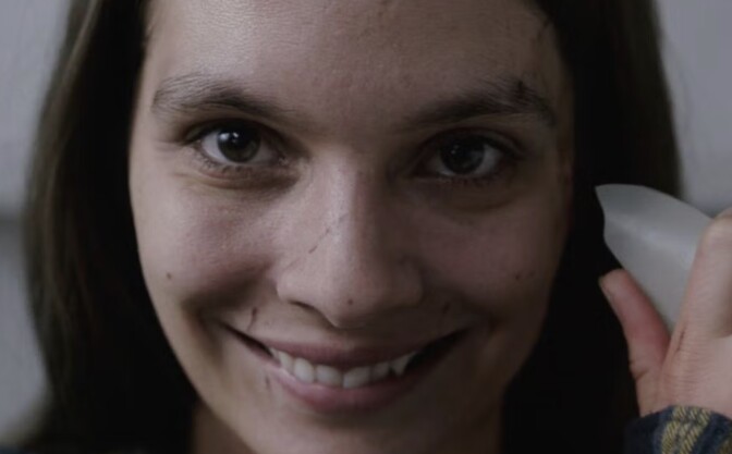 A photo of actress Caitlin Stasey in the horror movie "Smile." She is holding a sharp pottery shard to her face, which is scarred across her nose, and smiling in a disturbing manner.