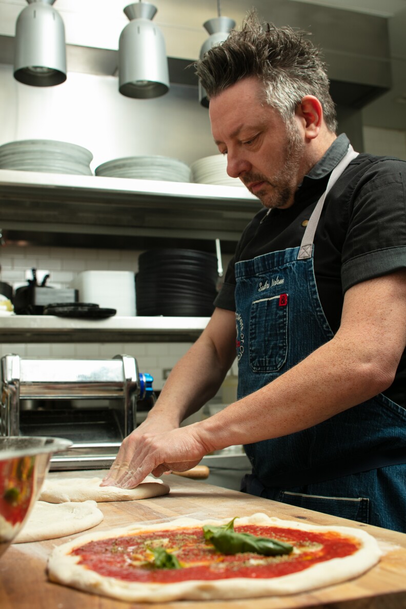 A man making pizza in a kitchen. A raw uncooked pizza with tomato sauce sits on a wooden surface, while the man who is wearing a dark denim apron kneads pizza dough next to it. Behind him are metal shelves containing a pasta maker and various plates and utensils.