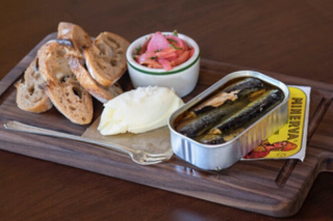 A wooden cutting board holds a pile of sliced bread with a small dish of pickled vegetables, a small amount of a buttery spread and a rectangular can of tinned fish.