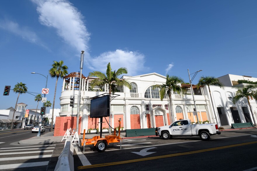 A white, elaborate storefront is shown on the corner of a street in Beverly Hills. A crosswalk is in the forefront and a white police pick-up truck is parked to the right.