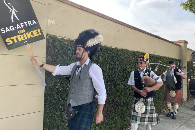 A bagpipe band marches in front of Warner Bros. studios in Burbank. They are in front of a beige wall with green ivy on it. The band wears kilts and traditional Scotiysh garb