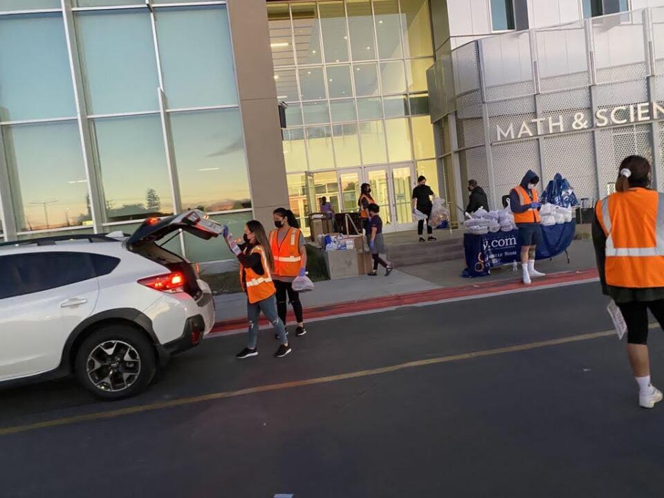Eight people, most wearing orange safety vests, work at a pickup line for meals. Two of the workers help load food into the back of a white station wagon.