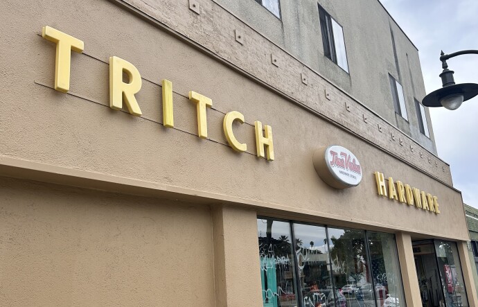 The exterior of a tan, two-story building on a partly-cloudy afternoon. The words "Tritch Hardware" are displayed across the front in yellow, with a smaller pop-out sign in between that reads "True Value" in red.