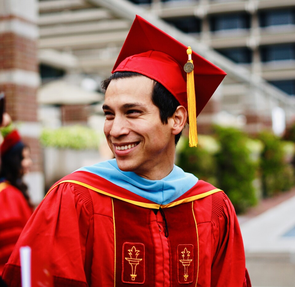 A man with short dark hair and medium-light skin tone smiles. He is clad in a cardinal red cap and gown with a light blue hood. 