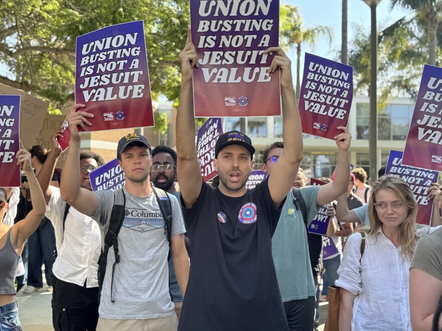 About two dozen people, with skin tones ranging from light to medium-dark, march on a college campuse while holding up signs that read: "UNION BUSTING IS NOT A JESUIT VALUE."