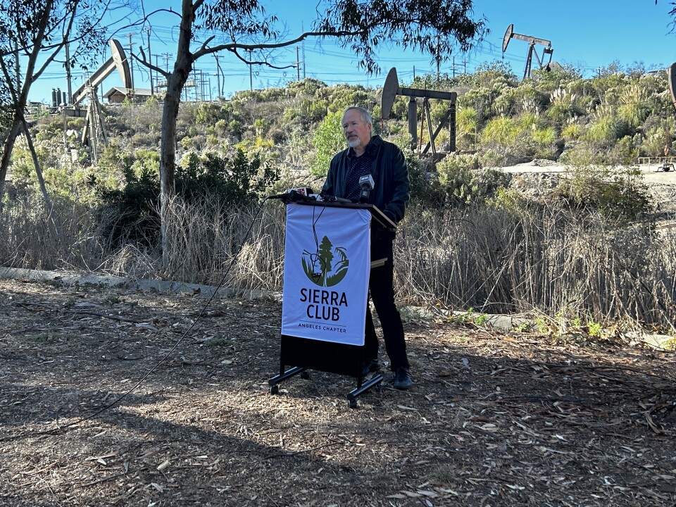 An older man with grey hair and light skin wearing a black long-sleeved shirt and pants stands at a podium with a sign reading "Sierra Club." In the background are oil pumpjacks. 