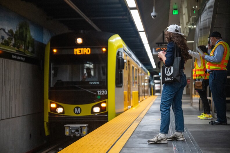 A black Metro subway train with yellow trim approaches the platform as three people wait. Two of them are wearing yellow and orange safety vests. The other is a woman wearing a white baseball cap with a black bag slung over one shoulder. The bag bears the word "Metro" and the organization's signature capital "M" in a white circle.