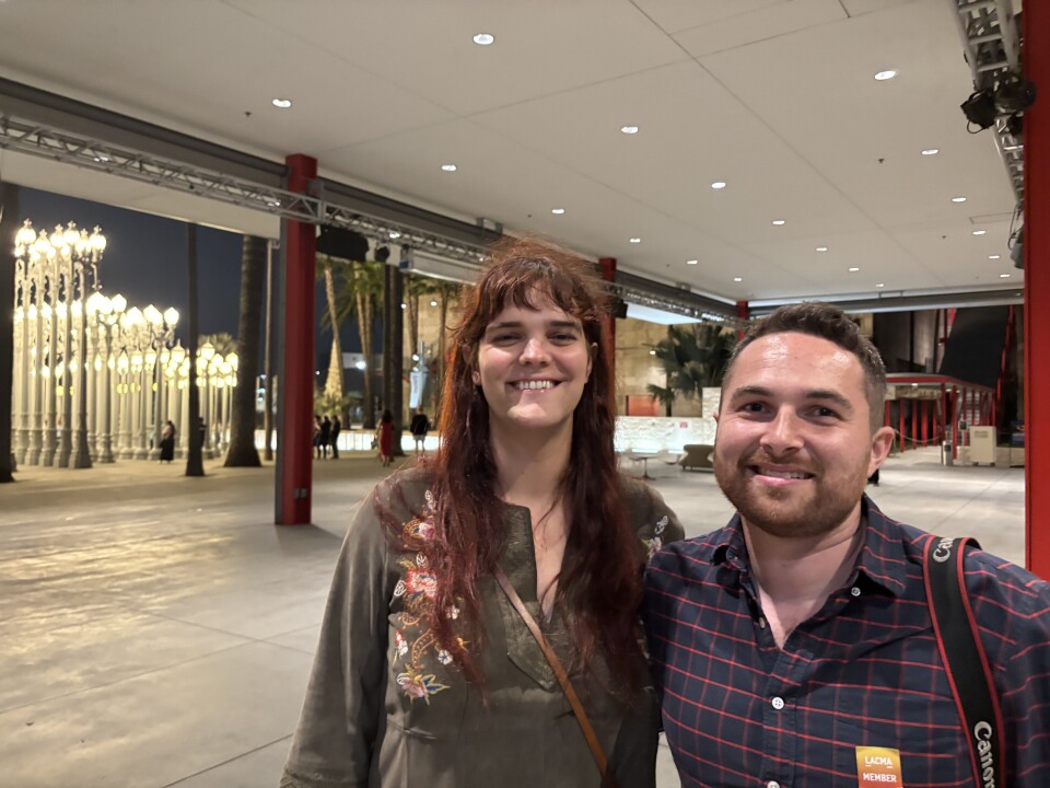 A woman with long red hair and a brown embroidered dress stands next to a man with a plaid shirt and camera over his shoulder. In the background are a collection of illuminated street lights, part of an outdoor art installation at LACMA.