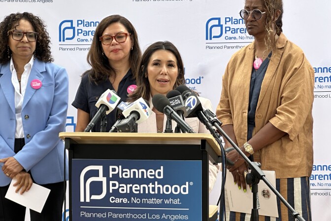 A group of four women stand at a podium during a Planned Parenthood Los Angeles press event. The woman at the podium with medium light skin tone speaks into multiple microphones from news outlets including ABC 7, KCAL News, and Univision. Behind her is a Planned Parenthood step-and-repeat banner. Two women wear pink “Bans Off Our Bodies” buttons. 