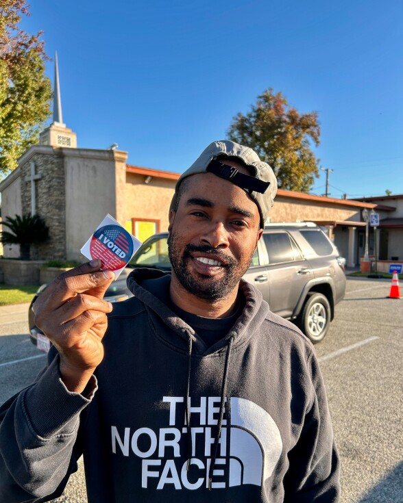 A man with medium-dark skin tone wearing a black The North Face sweater and gray cap worn backwards holds up an "I Voted" sticker. 