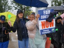 Two people stand under an umbrella smiling. One of them holds a sign that reads "Fair Contract NOW." One person with long brown hair and medium-light skin tone wears a blue hat. The other person with medium-dark skin tone wears a pink jacket and poncho.