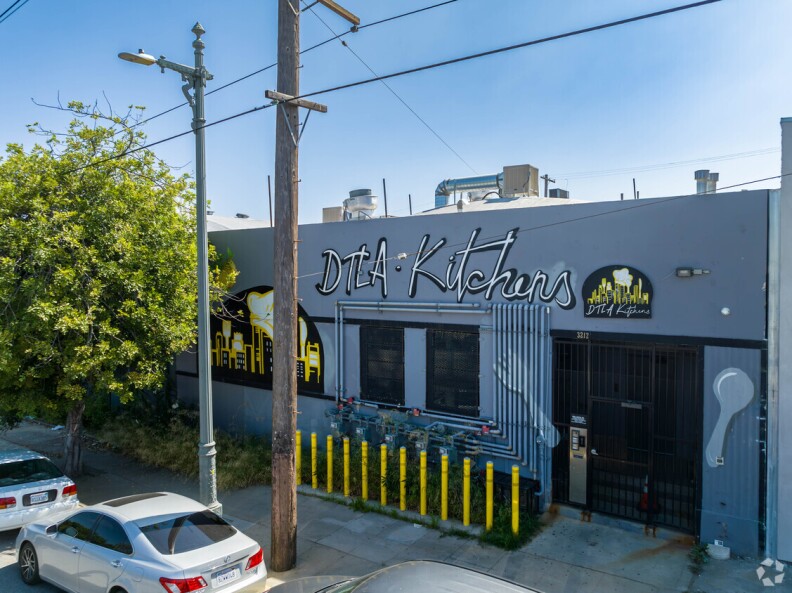 A grey building outside with a blue sky above. The building has black and white lettering that say "DTLA Kitchens" across the top, with black and yellow accenting. In front of the building is a black telephone wire and two cars parked along the sidewalk.