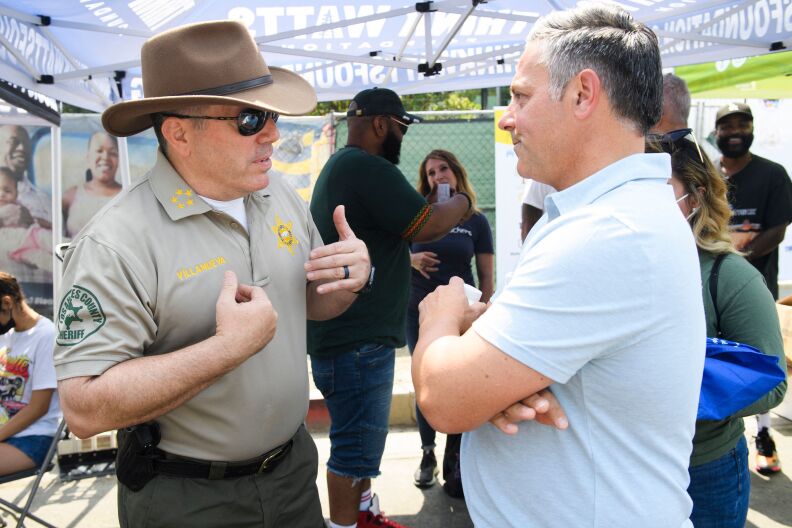 Sheriff Alex Villanueva wears a cowboy hat and stands talking to City Council member Joe Buscaino, who's in a polo shirt.