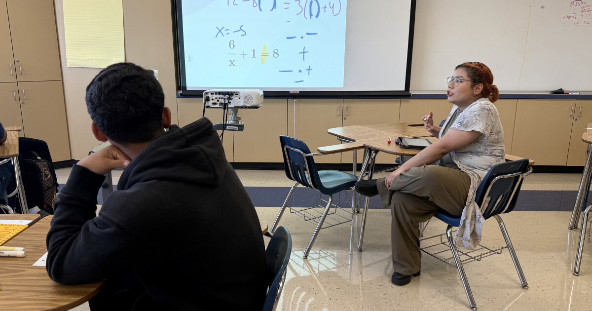 A teenage boy with medium skin tone and a black hoodie looks at a projection of math calculations. A women with medium light skin tone and glasses sits at a desk and gestures as though mid-speech.