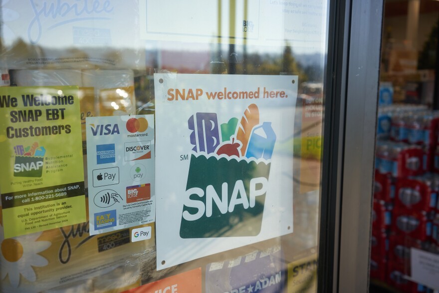 Close up of a glass door at a grocery store. Several signs hang on the door including a white sign with an illustration of a green grocery bag with a carton of eggs, broccoli, bread, milk and an apple. On the bag in white letters is the word "SNAP"
