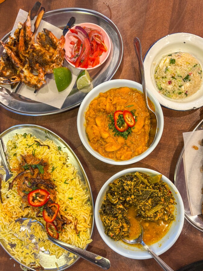 A colorful spread of East African dishes served on a wooden table. The meal includes skewered grilled prawns with lime wedges and sliced tomato-onion salad, a bowl of creamy bean stew topped with sliced red chili, a serving of yellow rice with caramelized onions and peppers, a leafy greens stew in a rich sauce, and a side of couscous-style grain mixed with vegetables.
