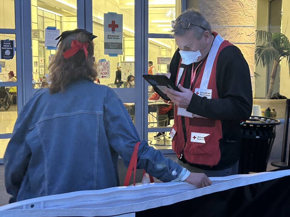 A man in red vest looks down at an iPad as a woman in a denim jacket looks on.