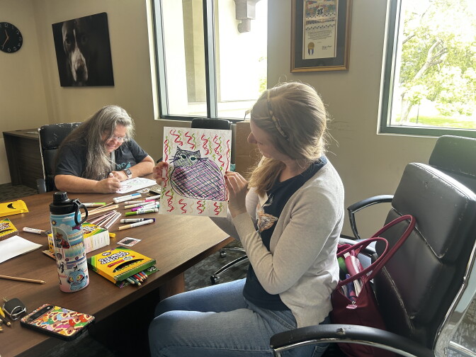 Two people at a table with colored pencils and markers, one holding up a drawing of an cat decorated with squiggly lines.