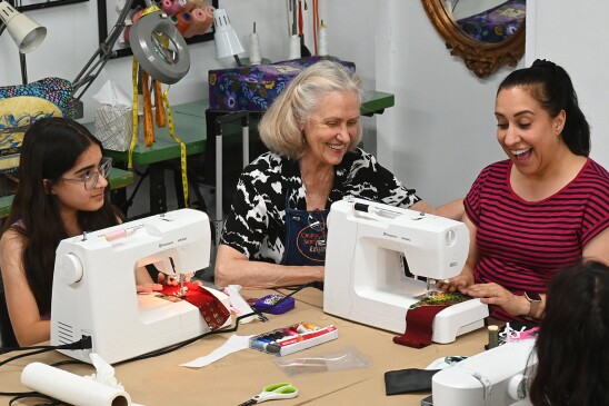 An older woman with graying hair smiles as she teaches three women sitting at sewing machines.