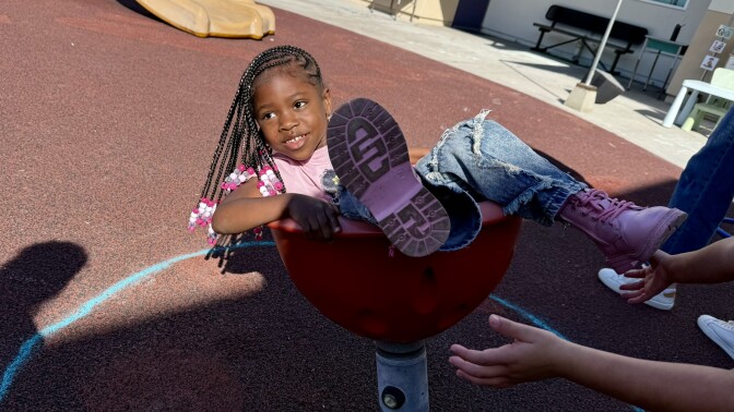  A student with dark skin tone and brown hair in braids with pink beads spins in a chair-like toy on the playground.