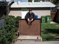 A rusty wide square door opens to a concrete base. A man stands in a yard holding it open. There's green grass and garbage cans behind him.