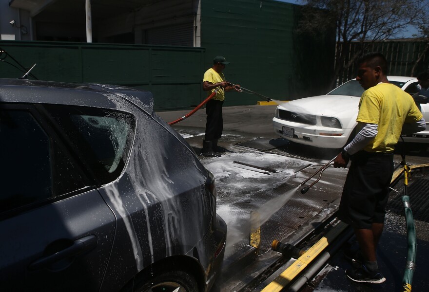 The back of a grey car is covered in suds as two workers, wearing yellow tops and black pants, wash off the foam using hoses