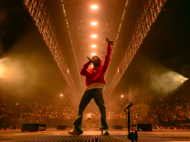 A man in a red sweatshirt stands on a stage with a mic in hand