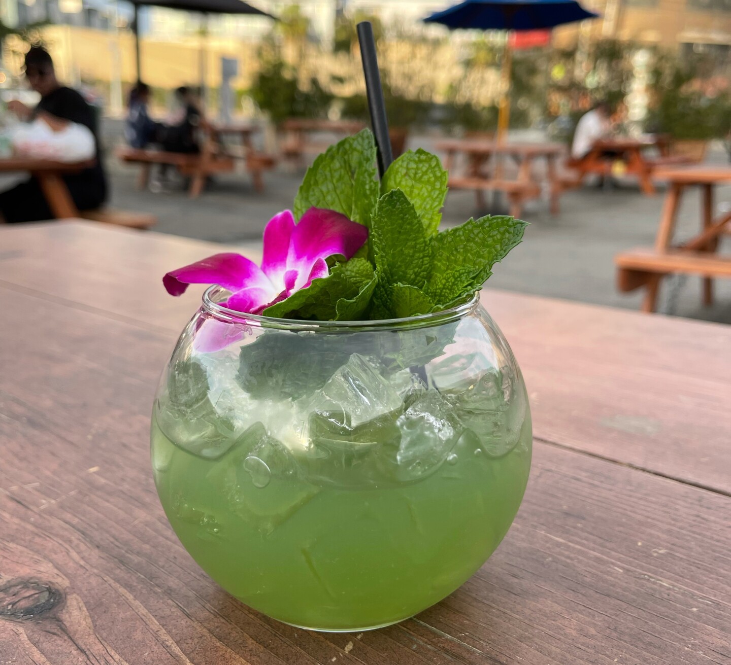 a glass bowl filled with green liquid and ice cubes and topped with mint leaves and a flower sits on a wooden table