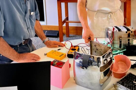 Two men stand at a table full of random gadgets and odd and ends. One is wearing a white apron and hat. 