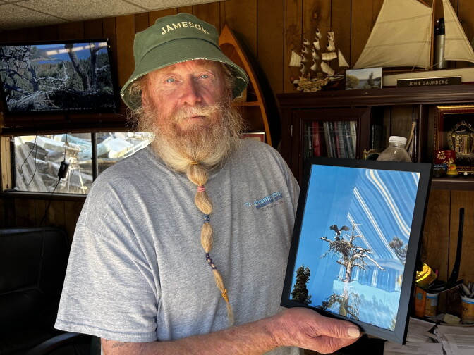 A bearded man in a green 'Jameson' bucket hat holds a framed photo of an eagle on a nest atop a tree.