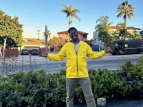 A Black man in a yellow jacket stands smiling in front of several rows of swiss chard planted in the ground with houses in the background