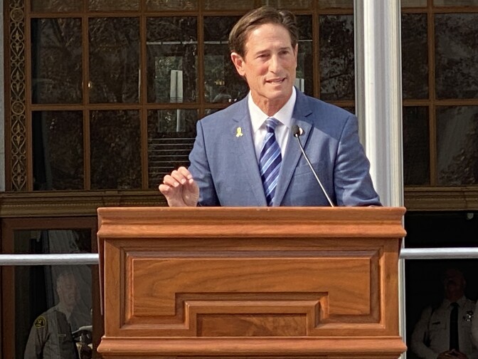 A white man in a dark blue suit stands alone outside during the day with a U.S. Flag in the background.