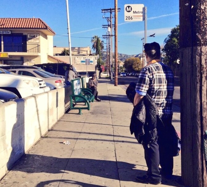 A man stands in the shadow cast by an electric pole as he waits for the bus at a stop with no bus shelter. Less than a quarter of L.A. bus stops have shelters and a city plan to add more has fallen apart.