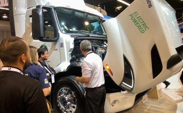 People gather around a large, white electric semi-truck that has it's hood open. 
