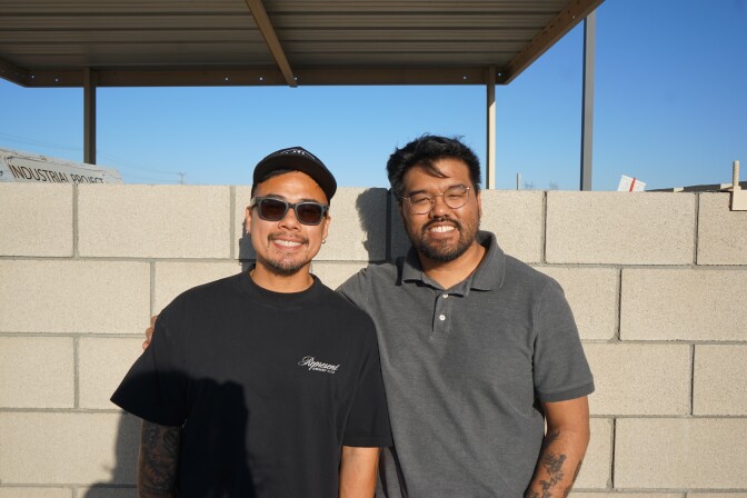 A medium-light skin-toned man with short hair wearing a black hat, shirt and sunglasses smiles at the camera. To his left, a second man with medium-light skin tone wearing glasses and a gray polo shirt also smiles. The man on the right has his arm around the man on the left.