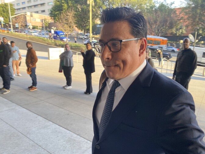 Jose Huizar, a brown-skinned man with black hair and glasses, is wearing a dark-colored suit and tie and a white shirt. He looks in the direction he is walking, to the left of the image and away from the camera