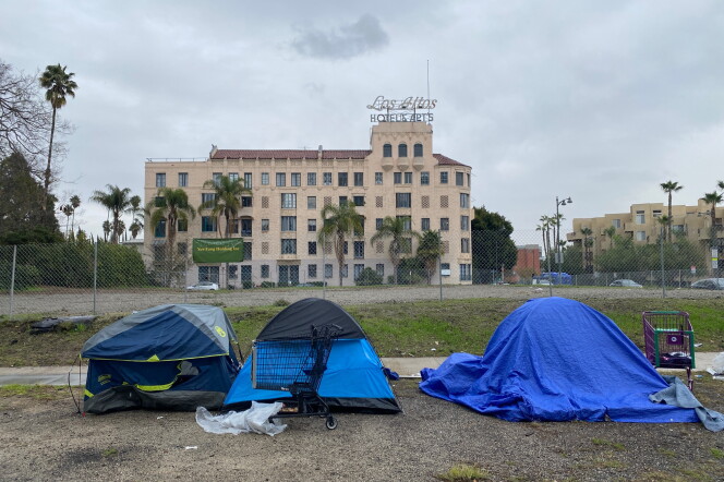 Three blue tents used by people experiencing homelessness are lined up on along a sidewalk. Behind them is a hotel.