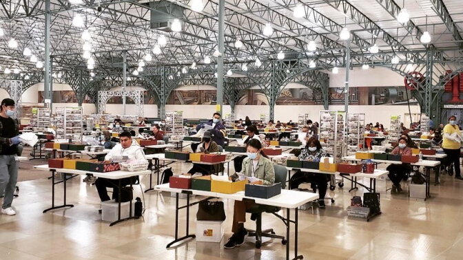 Election workers in a warehouse sit at voting tables and process votes