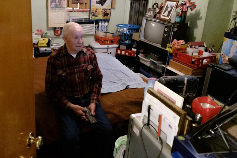 A man in plaid shirt sits on a bed in a small bedroom. A calendar, a pinup photo and other pictures hang on the wall behind him. A TV, laundry basket, mail and other personal effects are visible. The door of the room is halfway open.