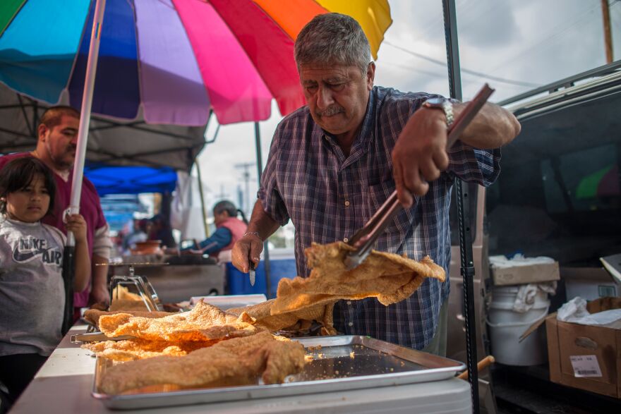 A man leans in to use tongs to pick up food on the top of his outdoor cart. Customers stand nearby and watch him.