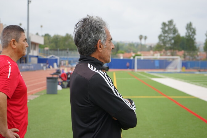 A man dressed in a black tracksuit looks off the frame and is turned slightly away with only a side profile of his face. He is standing on a green soccer pitch with a goal in the distance. Another person is standing beside him facing off the frame as well. 