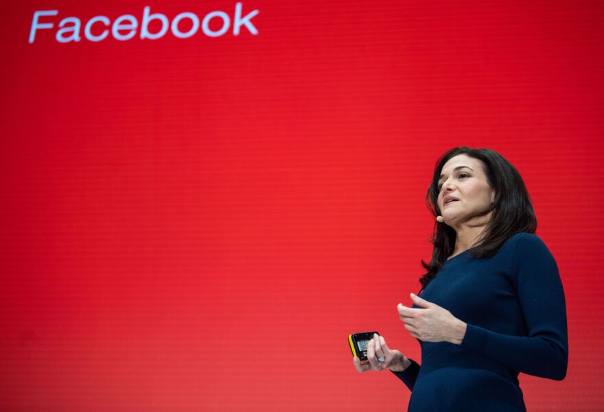 Sheryl Sandberg stands in a navy dress in front of a bright red background with the word Facebook at the top left in white