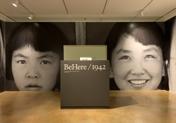 Photo of a room in a museum with giant photos of the faces of two young Japanese American girls plastered on one wall. In the foreground is a sign that reads "BeHere/1942," the name of the exhibition.