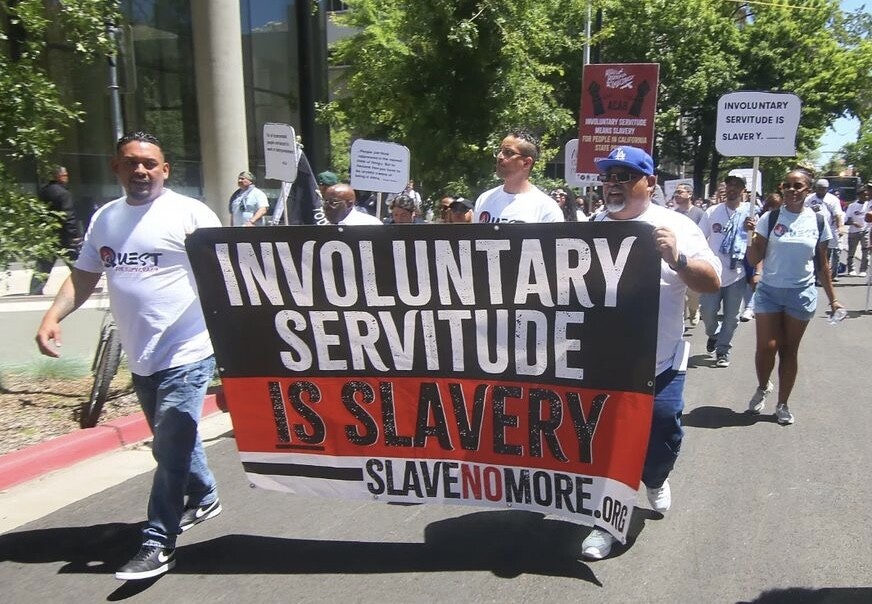 A crowd of people in white T-shirts march behind a banner sign that states "Involuntary Servitude Is Slavery."