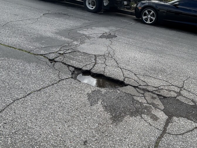 A close up of a street with a cracked pothole in the middle, which is full of rain water.