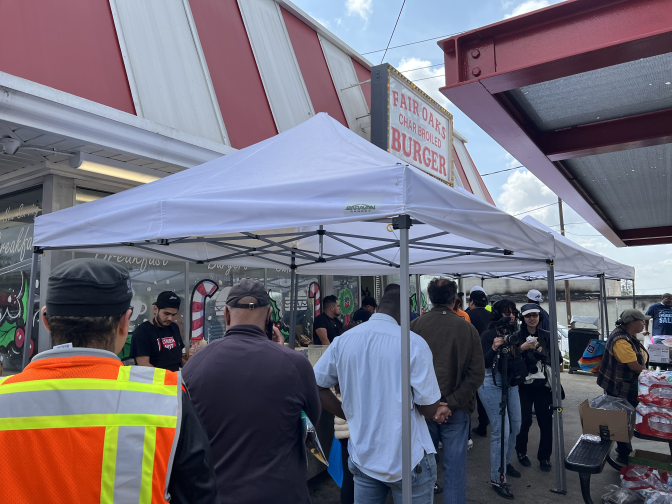 People stand under a white shade pop-up tent near a red and white roofed burger restaurant. 