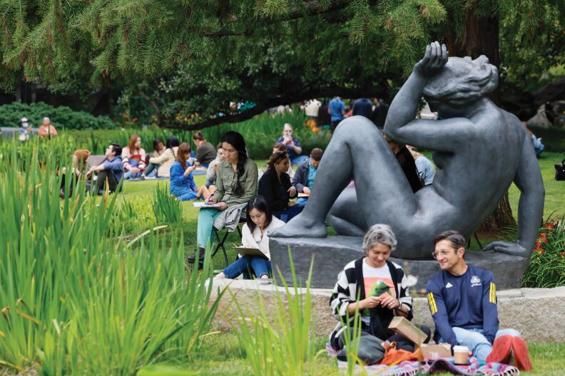 Groups of adults sit outside on picnic blankets and chairs among grass, plants, and trees. To the center left of the photo is a statue of a nude woman, with one patron of the park sitting at her feet, reading something on a clip board.